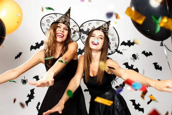 Zwei schöne Mädchen in schwarzen Kleidern und Hexenhüten haben Spaß mit Luftballons und Konfetti auf dem Hintergrund der Wand mit Fledermäusen — Stockfoto
