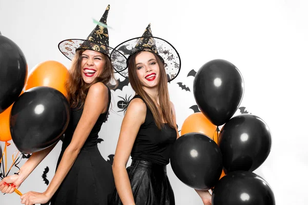 Dos hermosas chicas en vestidos negros y sombreros de brujas se divierten con globos negros y naranjas. Halloween — Foto de Stock