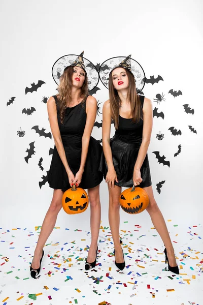Due bellissime ragazze che indossano abiti neri, cappelli da strega e tacchi alti tengono zucche di Halloween sullo sfondo con i pipistrelli — Foto Stock