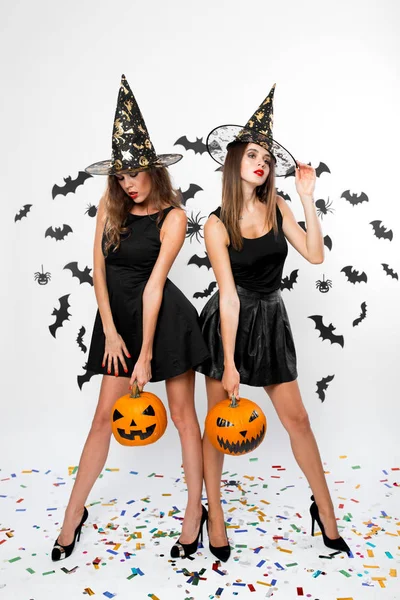 Due ragazze brune che indossano abiti neri, cappelli da strega e tacchi alti tengono zucche di Halloween sullo sfondo con i pipistrelli — Foto Stock