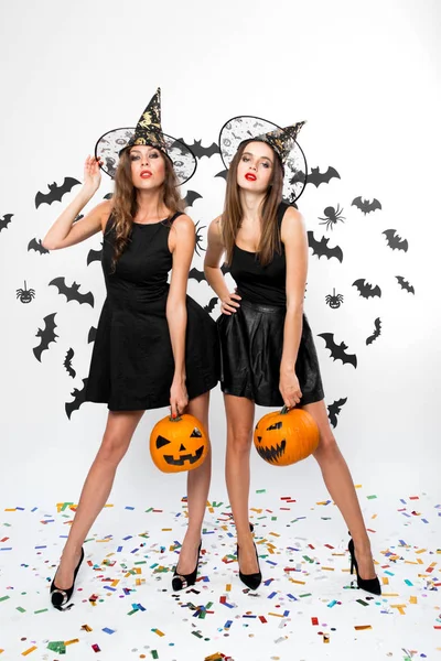 Zwei brünette Mädchen in schwarzen Kleidern, Hexenhüten und High Heels halten im Hintergrund Halloween-Kürbisse mit Fledermäusen — Stockfoto