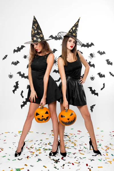 Due ragazze brune che indossano abiti neri, cappelli da strega e tacchi alti tengono zucche di Halloween sullo sfondo con i pipistrelli — Foto Stock