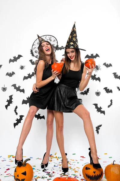 Zwei hinreißende Mädchen in schwarzen Kleidern, Hexenhüten und High Heels haben Spaß mit Halloween-Kürbissen — Stockfoto