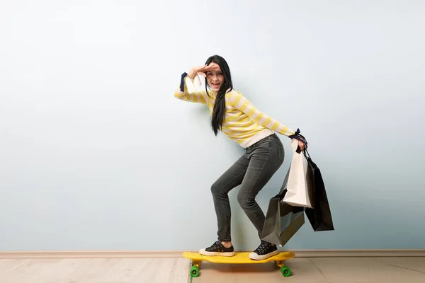 Joven chica morena vestida con blusa blanca y amarilla y jeans grises está en la tabla de skate amarilla sosteniendo un montón de bolsas después de comprar en el fondo blanco — Foto de Stock