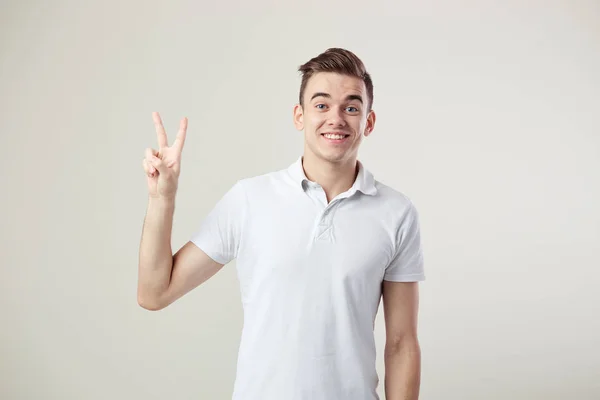 Affable  guy dressed in a white t-shirt and jeans shows  V sign on a white background in the studio — Stock Photo, Image