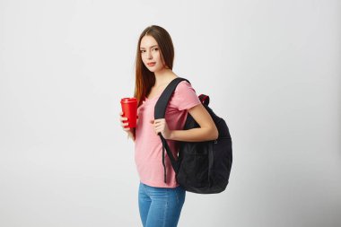 Omzuna siyah bir sırt çantası ile güzel kız pembe bir t-shirt giymiş ve kot kırmızı plastik bir kap beyaz zemin üzerine onun elinde tutan
