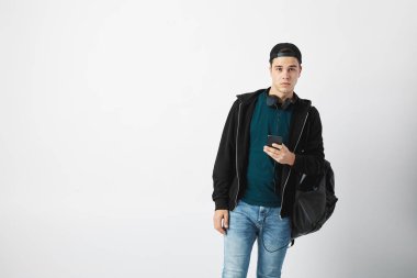 Karanlık bir t-shirt, kot pantolon, kazak ve bir kap kullandığı cep telefonu Studio beyaz bir arka plan üzerinde siyah bir sırt çantası onun omuz ve kulaklık onun boynuna adamla giymiş