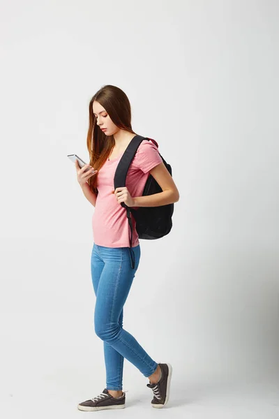 Trevlig tjej med en svart ryggsäck på hennes axel klädd i en rosa t-shirt, jeans och sneakers använder mobiltelefon på en vit bakgrund — Stockfoto