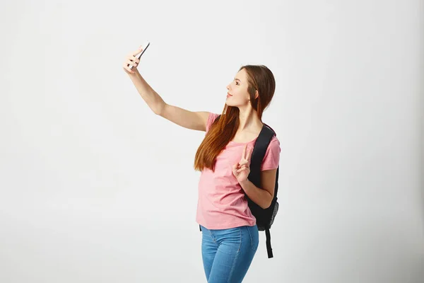 Linda chica con una mochila negra en el hombro vestida con una camiseta rosa y jeans hace una selfie sobre un fondo blanco — Foto de Stock