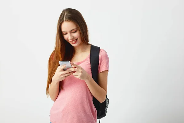 Linda chica con una mochila negra en el hombro vestida con una camiseta rosa utiliza el teléfono móvil sobre un fondo blanco — Foto de Stock