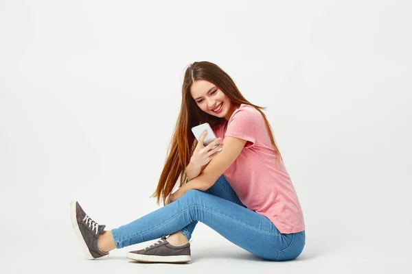 Hermosa chica vestida con una camiseta rosa, jeans y zapatillas de deporte se sienta en el suelo y utiliza el teléfono móvil sobre un fondo blanco en el estudio — Foto de Stock