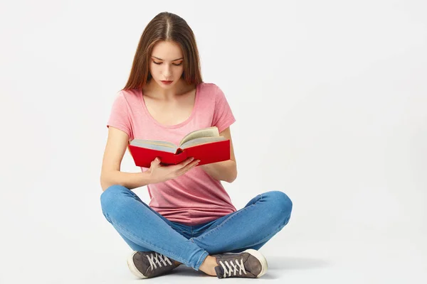 Belle fille vêtue d'un t-shirt rose, jeans et baskets s'assoit sur le sol et lit un livre sur fond blanc dans le studio — Photo