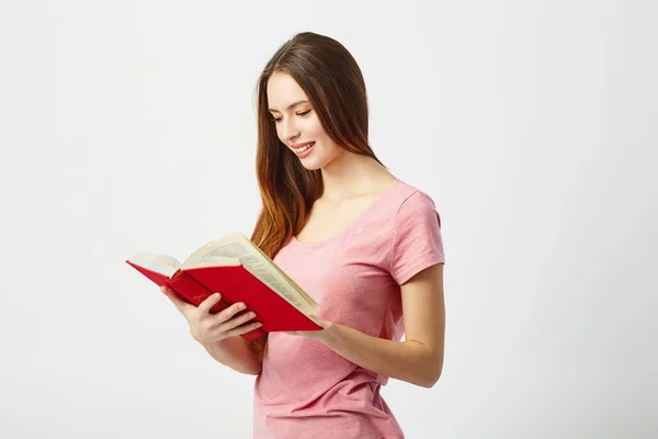 Belle fille aux cheveux longs vêtue d'un t-shirt rose lit un livre sur fond blanc en studio — Photo