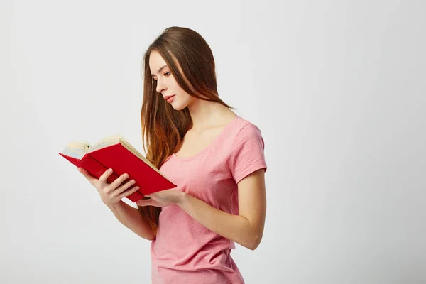 Красивая длинноволосая девушка в розовой футболке читает книгу на белом фоне в студии — стоковое фото