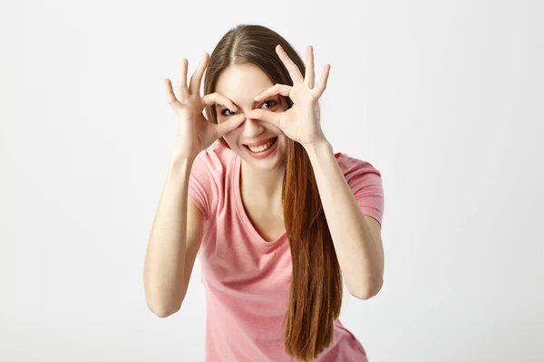 Grappig langharige meisje gekleed in een roze t-shirt organiseert haar handen in de vorm van bril over haar ogen op een witte achtergrond in de studio — Stockfoto