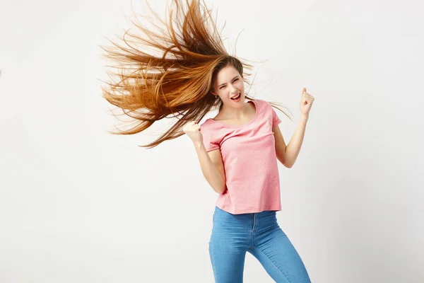 Ragazza divertente con i capelli lunghi svolazzando nel vento vestita una t-shirt rosa e jeans si diverte su uno sfondo bianco in studio — Foto Stock