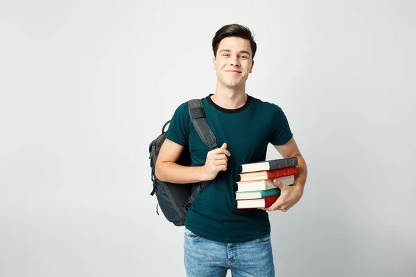 Улыбающийся темноволосый парень с черным рюкзаком на плече, одетый в черную футболку и джинсы держит книги в руках на белом фоне — стоковое фото