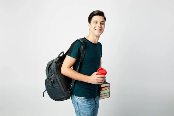 Leende mörkhårig kille med en svart ryggsäck på hans axel klädd i en svart t-shirt och jeans håller böcker och en röda plastmugg i händerna på en vit bakgrund — Stockfoto