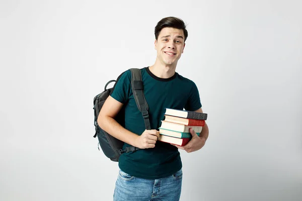Mörkhårig kille med en svart ryggsäck på hans axel klädd i en mörk t-shirt och jeans håller böcker i händerna på en vit bakgrund — Stockfoto