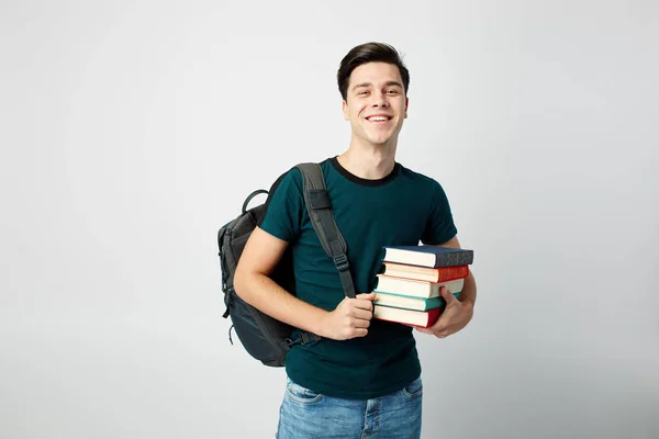Dark-haired guy with a black backpack on his shoulder dressed in a dark t-shirt and jeans holds books in his hands on a white background