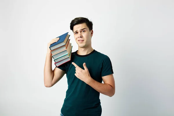 Engraçado cara de cabelos escuros vestido com uma camiseta escura e jeans mantém livros em seu ombro em um fundo branco — Fotografia de Stock