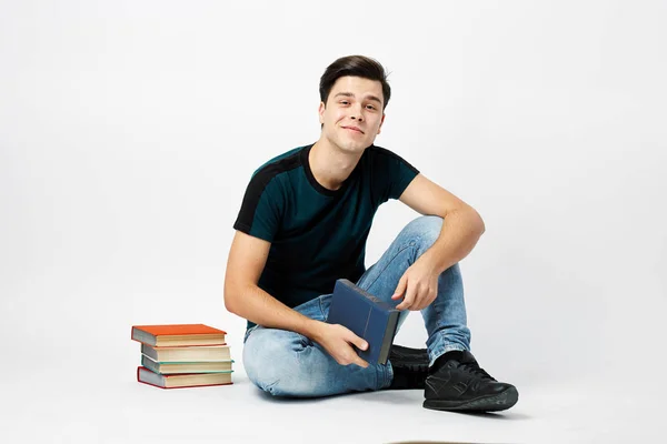 Bonito cara de cabelos escuros vestido com uma camiseta escura e jeans senta-se no chão e lê um livro sobre um fundo branco no estúdio — Fotografia de Stock