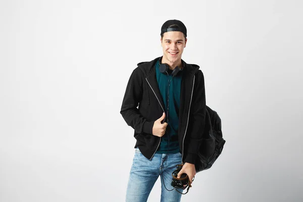 Un gars souriant avec un sac à dos noir sur l'épaule habillé d'un t-shirt vert foncé, un jean, un sweat-shirt et une casquette tient une caméra dans sa main sur un fond blanc — Photo