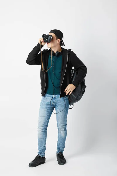 Le gars avec un sac à dos noir sur l'épaule habillé d'un t-shirt foncé, un jean, un sweat-shirt et une casquette fait une photo en studio sur un fond blanc — Photo