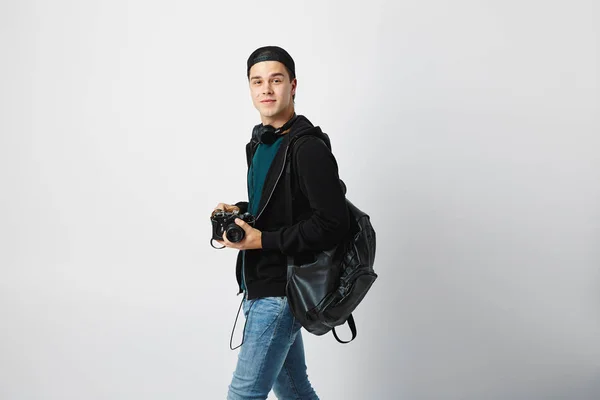 Sorrindo cara com uma mochila preta em seu ombro vestido com uma camiseta verde escuro, jeans, camisola e um boné segura uma câmera em sua mão em um fundo branco — Fotografia de Stock