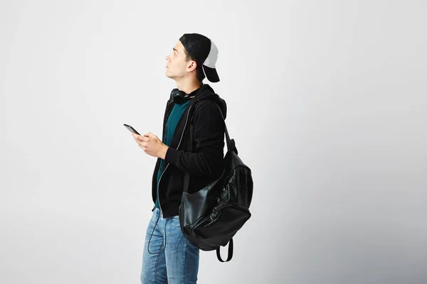 Le gars avec un sac à dos noir sur son épaule et des écouteurs autour de son cou habillé d'un t-shirt foncé, un jean, un sweat-shirt et une casquette utilise un téléphone portable dans le studio sur un fond blanc — Photo