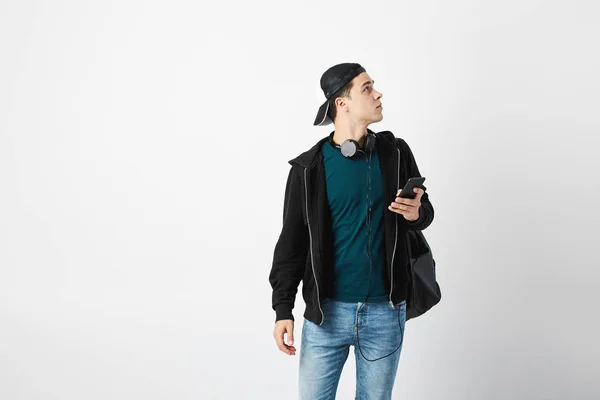 Le gars avec un sac à dos noir sur son épaule et des écouteurs autour de son cou habillé d'un t-shirt foncé, un jean, un sweat-shirt et une casquette utilise un téléphone portable dans le studio sur un fond blanc — Photo