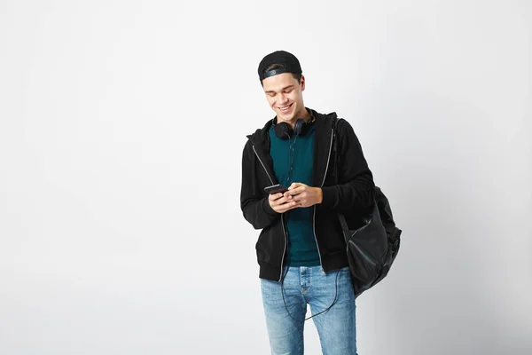 Kille med en svart ryggsäck på hans axel och hörlurarna runt halsen klädd i en mörk t-shirt, jeans, tröja och en mössa använder mobiltelefon i studion på en vit bakgrund — Stockfoto