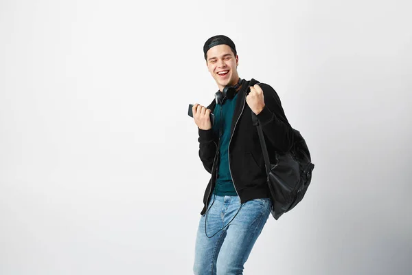 Šťastný chlap s černý batoh na rameno a sluchátka na hlavě na sobě tmavé tričko, džíny, mikinu a čepici používá mobilní telefon v ateliéru na bílém pozadí — Stock fotografie