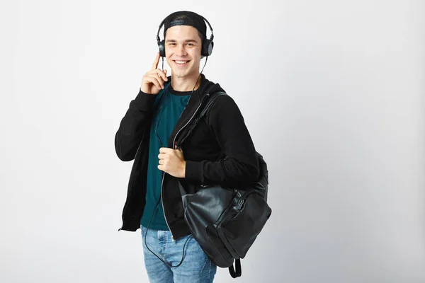 Heureux mec avec un sac à dos noir sur son épaule et écouteurs sur sa tête habillé d'un t-shirt sombre, jeans, sweat-shirt et une casquette utilise téléphone portable dans le studio sur un fond blanc — Photo