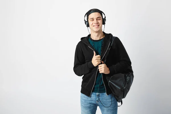 Glad kille med en svart ryggsäck på hans axel och hörlurar på huvudet klädd i en mörk t-shirt, jeans, tröja och en mössa använder mobiltelefon i studion på en vit bakgrund — Stockfoto