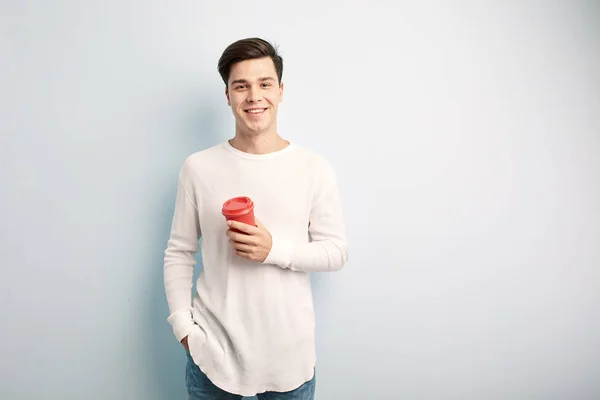 Cara de cabelos escuros vestida com uma camiseta branca de manga comprida e jeans segura um copo de plástico vermelho em sua mão em um fundo branco — Fotografia de Stock