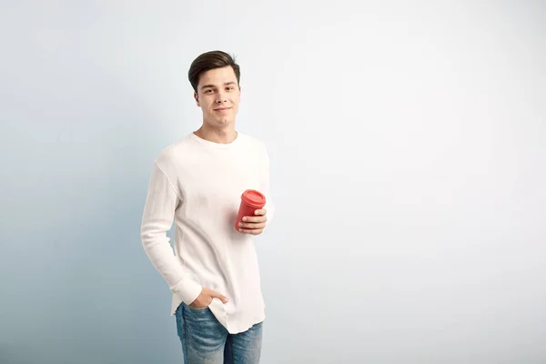 Cara de cabelos escuros vestida com uma camiseta branca de manga comprida e jeans segura um copo de plástico vermelho em sua mão em um fundo branco — Fotografia de Stock