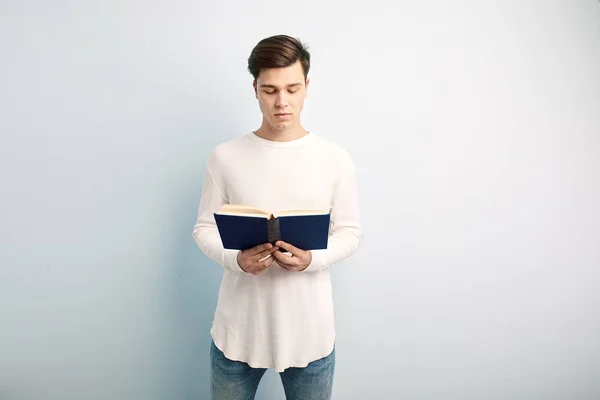 Homme aux cheveux bruns vêtu d'un t-shirt blanc à manches longues et un jean lit un livre dans sa main sur un fond blanc — Photo