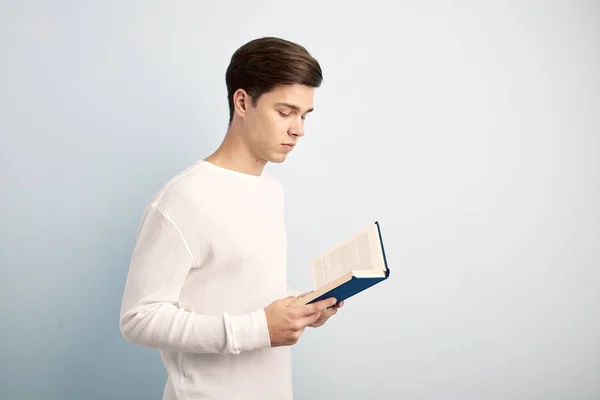 Dark-haired guy dressed in a white long sleeve t-shirt and jeans reads a book in his hand on a white background