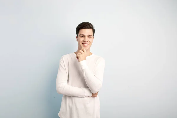 Sorrindo cara de cabelos escuros vestido com uma t-shirt manga longa branca e jeans fica e mantém a mão em seu queixo em um fundo branco — Fotografia de Stock