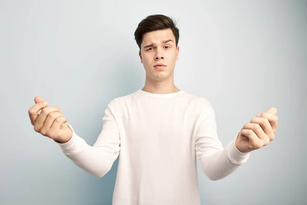 Engraçado cara de cabelos escuros vestido com uma t-shirt manga comprida branca e jeans fica e mantém as mãos na frente dele em um fundo branco — Fotografia de Stock