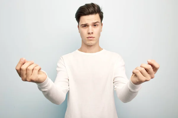 Engraçado cara de cabelos escuros vestido com uma t-shirt manga comprida branca e jeans fica e mantém as mãos na frente dele em um fundo branco — Fotografia de Stock