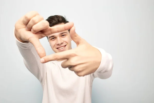 Engraçado cara de cabelos escuros vestido com uma t-shirt manga comprida branca fica e mantém as mãos na frente dele fazendo com os dedos uma forma de uma moldura em um fundo branco — Fotografia de Stock