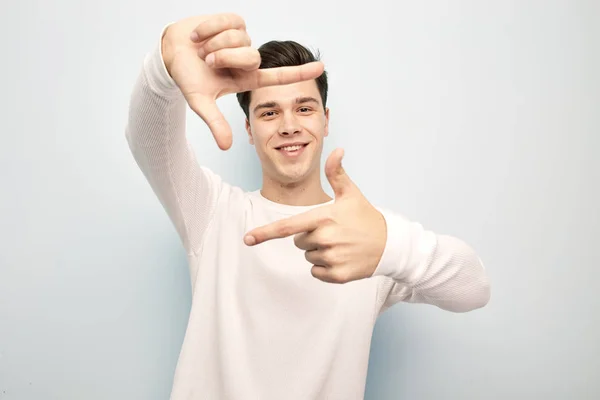 Rolig mörkhårig kille klädd i en vit långärmad t-shirt står och håller händerna framför honom att göra en form av en ram på en vit bakgrund med fingrar — Stockfoto