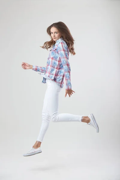 Chica alegre vestida con una camisa a cuadros y jeans blancos salta sobre el fondo blanco en el estudio — Foto de Stock
