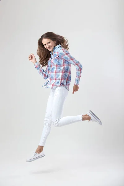 Chica alegre vestida con una camisa a cuadros y jeans blancos salta sobre el fondo blanco en el estudio — Foto de Stock