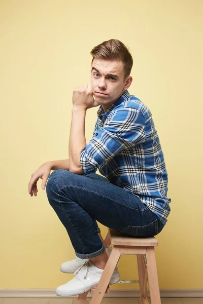 Ragazzo contemplativo vestito con una camicia a quadri e jeans è seduto su uno sgabello su uno sfondo giallo in studio — Foto Stock