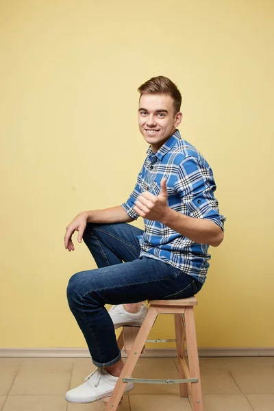 Ragazzo allegro vestito con una camicia a quadri e jeans è seduto e dare pollici su uno sgabello su uno sfondo giallo in studio — Foto Stock