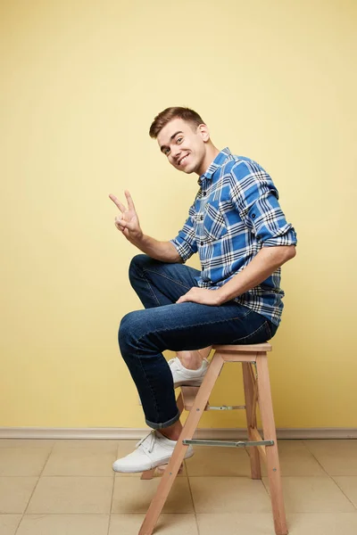 Glad kille klädd i en rutig skjorta och jeans sitter på en pall och visar V-tecknet på en gul bakgrund i studion — Stockfoto