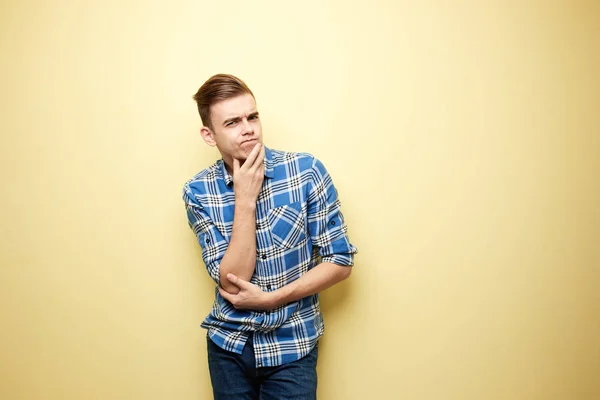 Pensivo l cara vestido com uma camisa xadrez e jeans fica ao lado da parede amarela no estúdio — Fotografia de Stock